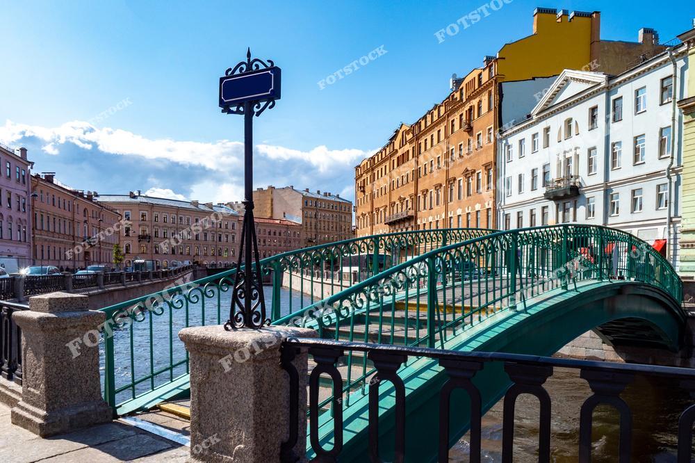На фотографии изображен Сенной мост, перекинутый через канал Грибоедова в Санкт-Петербурге. Этот пешеходный мост с изящными коваными перилами соединяет улицы исторического центра и создает уютное пространство для прогулок. Окружающие здания в пастельных тонах добавляют атмосферу старого Петербурга. Спокойные воды канала Грибоедова и ясное небо подчеркивают красоту летнего дня в городе.